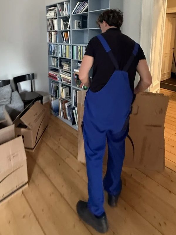 Bücher vom Regal in den Karton Ein Umzugshelfer in blauer Latzhose trägt einen Umzugskarton an einem vollen Bücherregal vorbei über den Dielenboden einer Altbauwohnung
