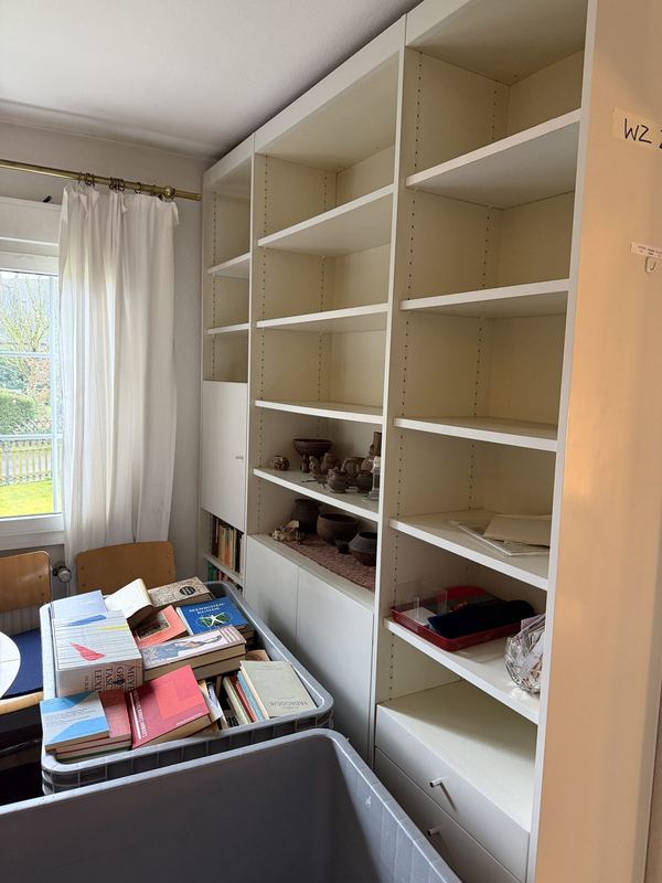 Fast leeres Bücherregal Ein weißes Regal, aus dem fast alle Bücher entfernt sind, vorne steht ein grauer Rollbehälter voller Bücher
