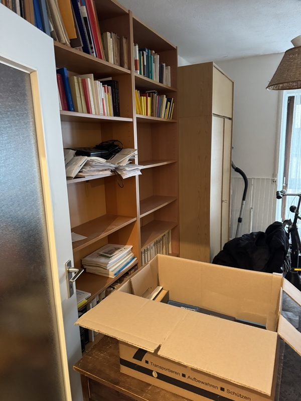 Ein aufgeklappter brauner Karton steht vor einem Regal mit Büchern, Ordnern und losen Blättern