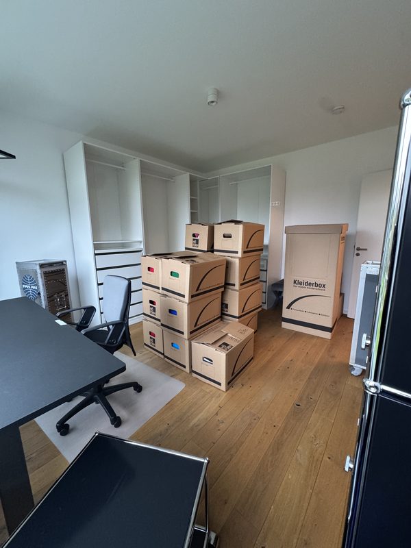 Gestapelte Umzugskartons, eine Kleiderbox und ein schwarzer Schreibtisch mit Bürostuhl in einem Raum mit Holzboden, bei einem Umzug in Leverkusen