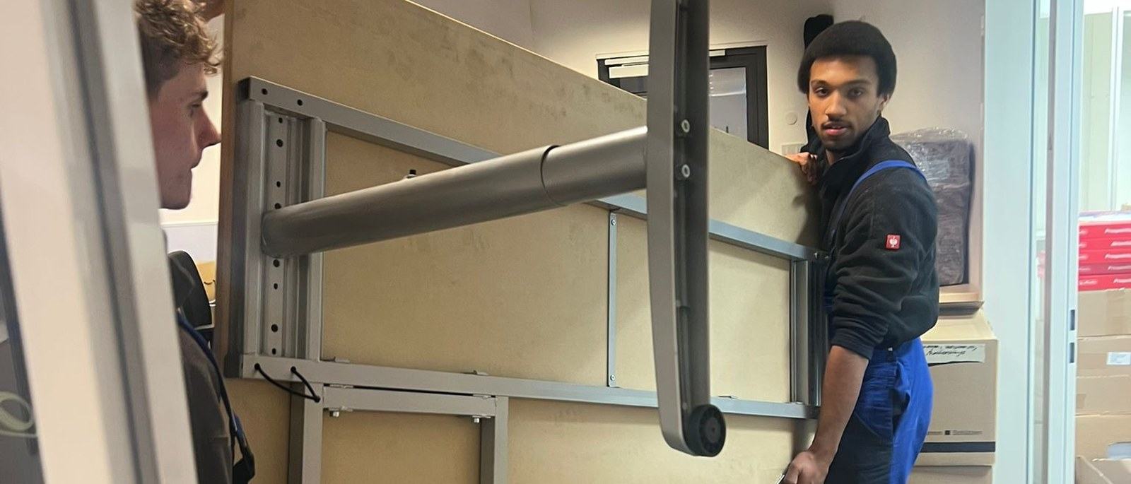 Büroumzug in Leverkusen: Ein hochkant gestellter Schreibtisch wird von zwei Mitarbeitern durch eine Bürotür getragen, im Hintergrund stapeln sich Kartons