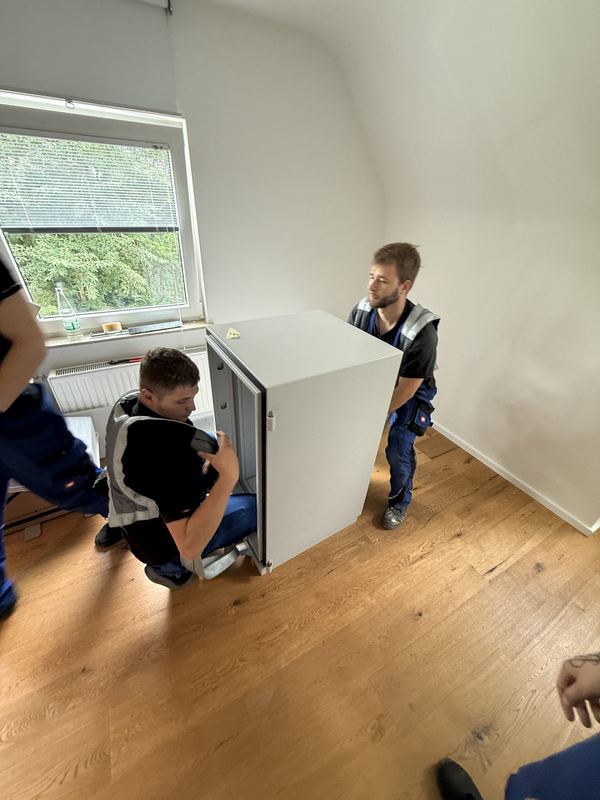 Zwei Kollegen mit einem weißen Einbaugerät auf dem Weg durch einen leergeräumten Raum