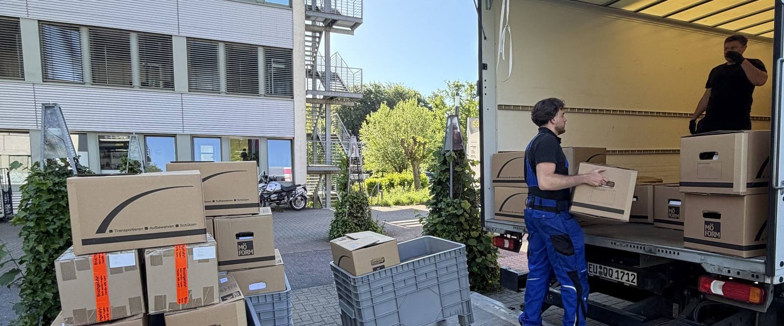 Einblicke in unseren Arbeitsalltag in Leverkusen: Vor einem Bürogebäude mit Außentreppe steht ein Umzugswagen mit offenem Laderaum, ein Mitarbeiter trägt einen Karton zur Ladebordwand, ein zweiter steht im Laderaum, im Vordergrund gestapelte Umzugskartons und graue Kunststoffboxen.