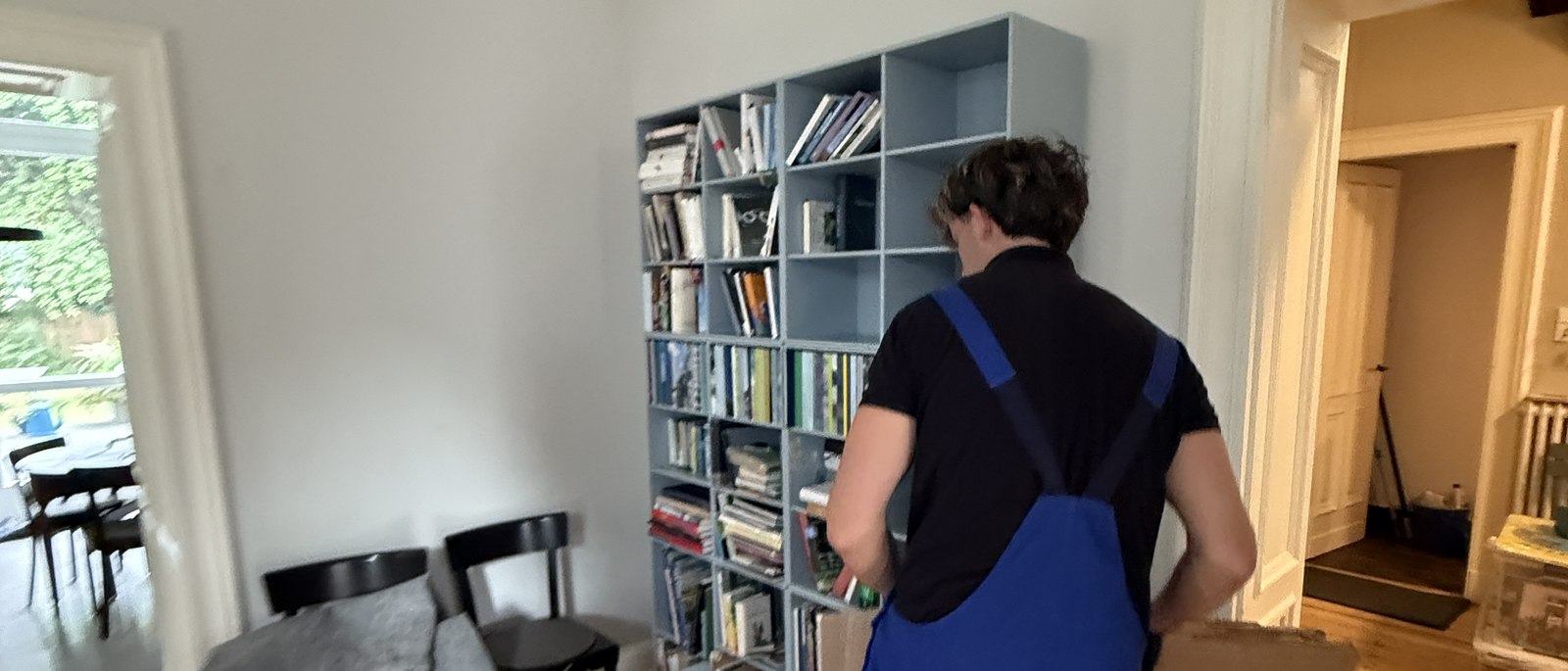 Fragen und Antworten zum Umzug in Leverkusen: Ein Mitarbeiter in Latzhose steht vor einem hellgrauen Würfelregal mit Büchern und Ordnern und packt dessen Inhalt in einen Karton, im Vordergrund ein Esstisch mit Stühlen.