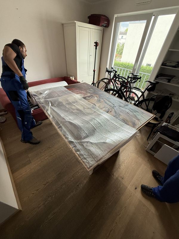 Rahmenbild in Luftpolsterfolie Großformatiges Bild im Rahmen, rundum in Luftpolsterfolie gewickelt, auf einem Tisch abgelegt, dahinter Fahrräder und ein Mitarbeiter