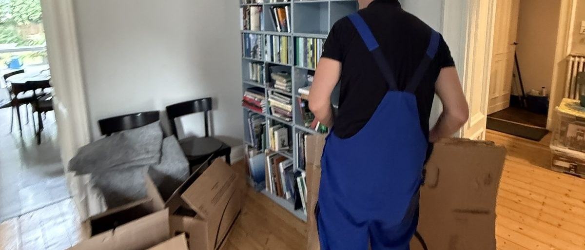 Haushaltsauflösung in Leverkusen: Ein Mitarbeiter in Latzhose geht mit einem gefalteten Karton an einem gut gefüllten Bücherregal vorbei, davor Stühle und aufgestapelte Kartons
