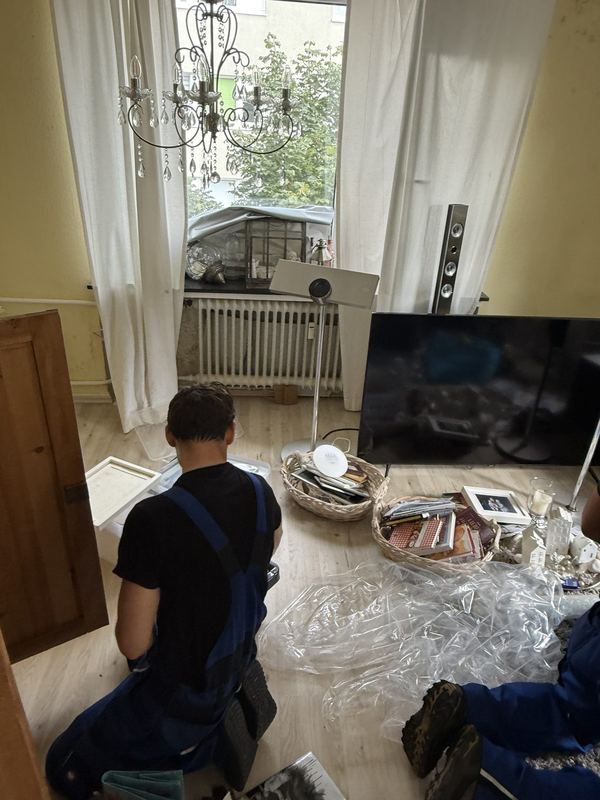 Hausrat durchsehen und aussortieren In einem Wohnzimmer kniet ein Mitarbeiter neben einem Korb voller Hausrat, am Boden ein Fernseher sowie Verpackungsfolie