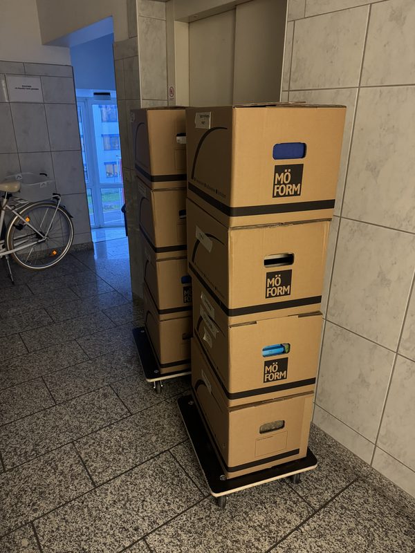 Mehrere Kartons übereinander auf einem Rollwagen, abgestellt in einem Hausflur mit Fliesen