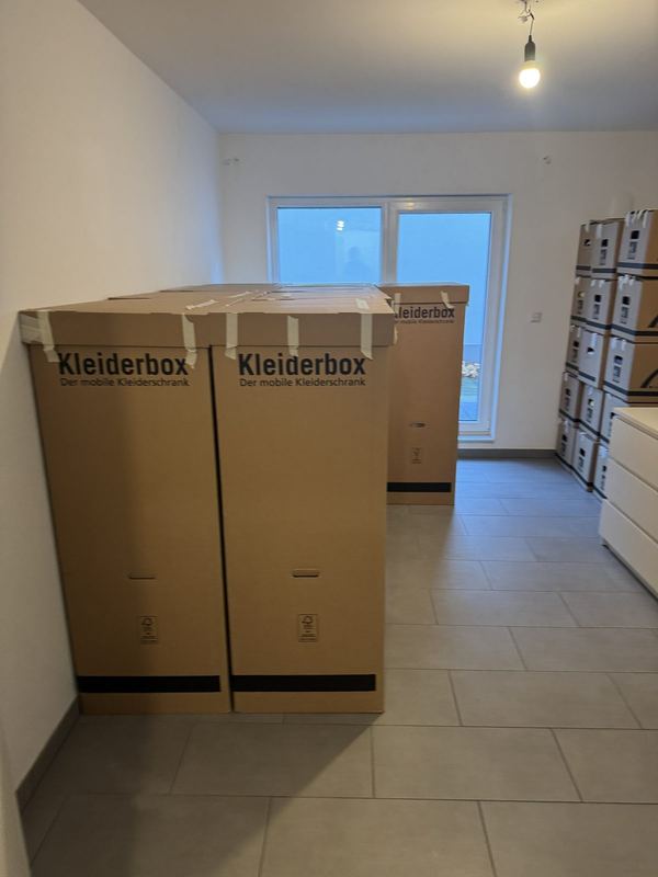 Zwei Kleiderboxen neben gestapelten Kartons in einem noch leeren Zimmer