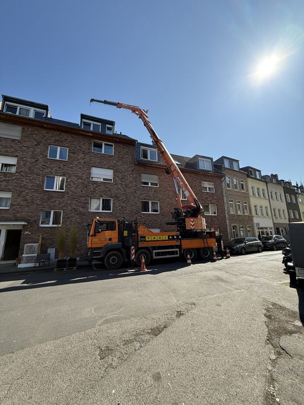 Orangefarbener Kranwagen in einer Wohnstraße, der Ausleger ist ausgefahren, Warnkegel stehen davor