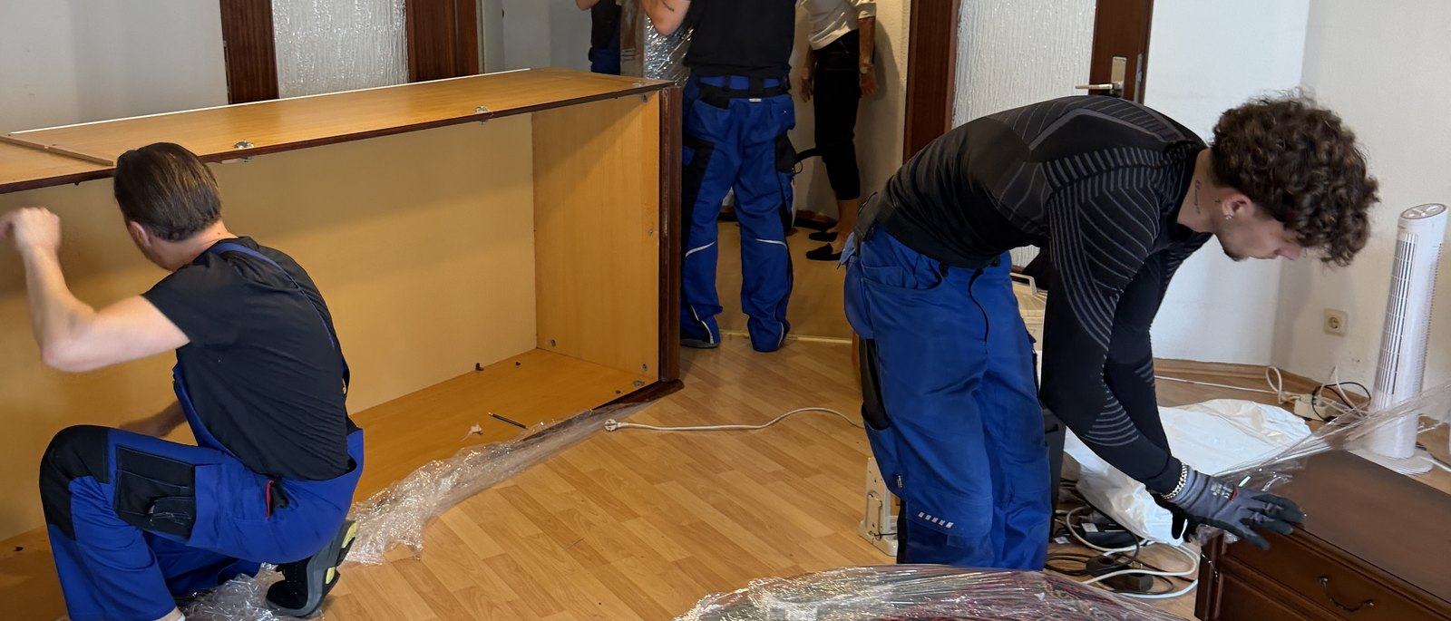 Möbelmontage in Leverkusen: Drei Mitarbeiter zerlegen in einem Wohnzimmer mit Parkett eine große Schrankwand aus Holz, die Seitenteile liegen bereits am Boden, daneben Folie und Werkzeug.