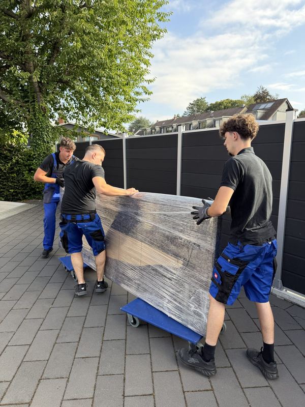 Drei Kollegen rollen ein in Folie gepacktes Möbelstück über eine Einfahrt, darunter Rollbretter
