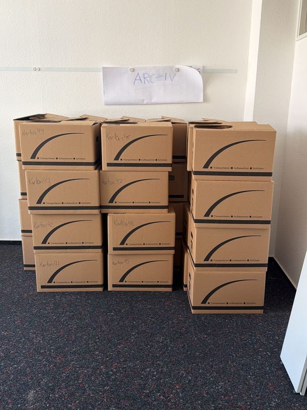 Ein handgeschriebenes Raumschild in einem geleerten Büro, darunter stapeln sich nummerierte Kartons
