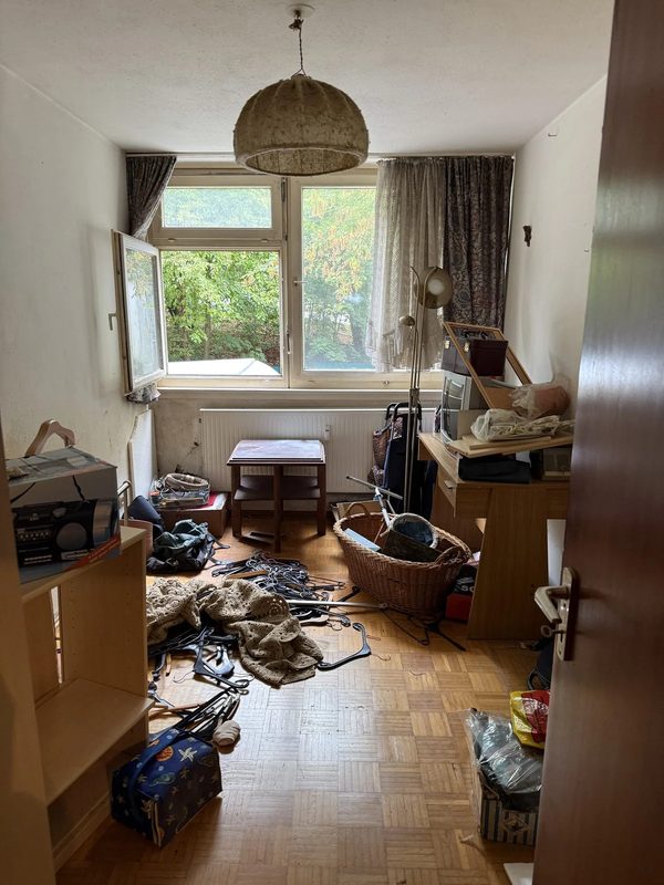 Raum vor der Entrümpelung Ein Zimmer mit Parkettboden vor der Entrümpelung: Kleiderbügel liegen verstreut auf dem Boden, dazu Kartons, ein Weidenkorb, eine Stehlampe und ein kleiner Tisch