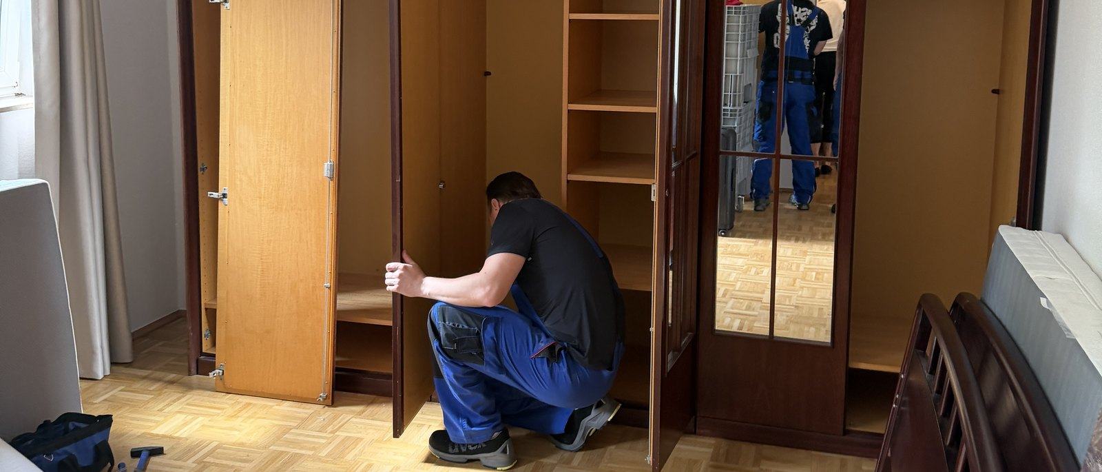 Seniorenumzug in Leverkusen: In einem Schlafzimmer mit Parkettboden kniet ein Mitarbeiter vor einem leer geräumten Kleiderschrank aus dunklem Holz
