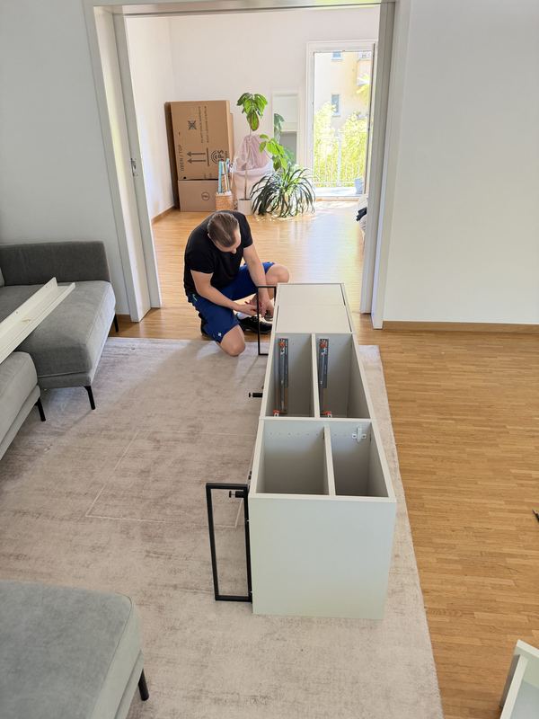 Sideboard-Montage in der neuen Wohnung in Leverkusen Ein Kollege kniet in einem hell eingerichteten Wohnzimmer vor einem weißen Sideboard und schraubt es zusammen, hinter ihm stapeln sich Umzugskartons