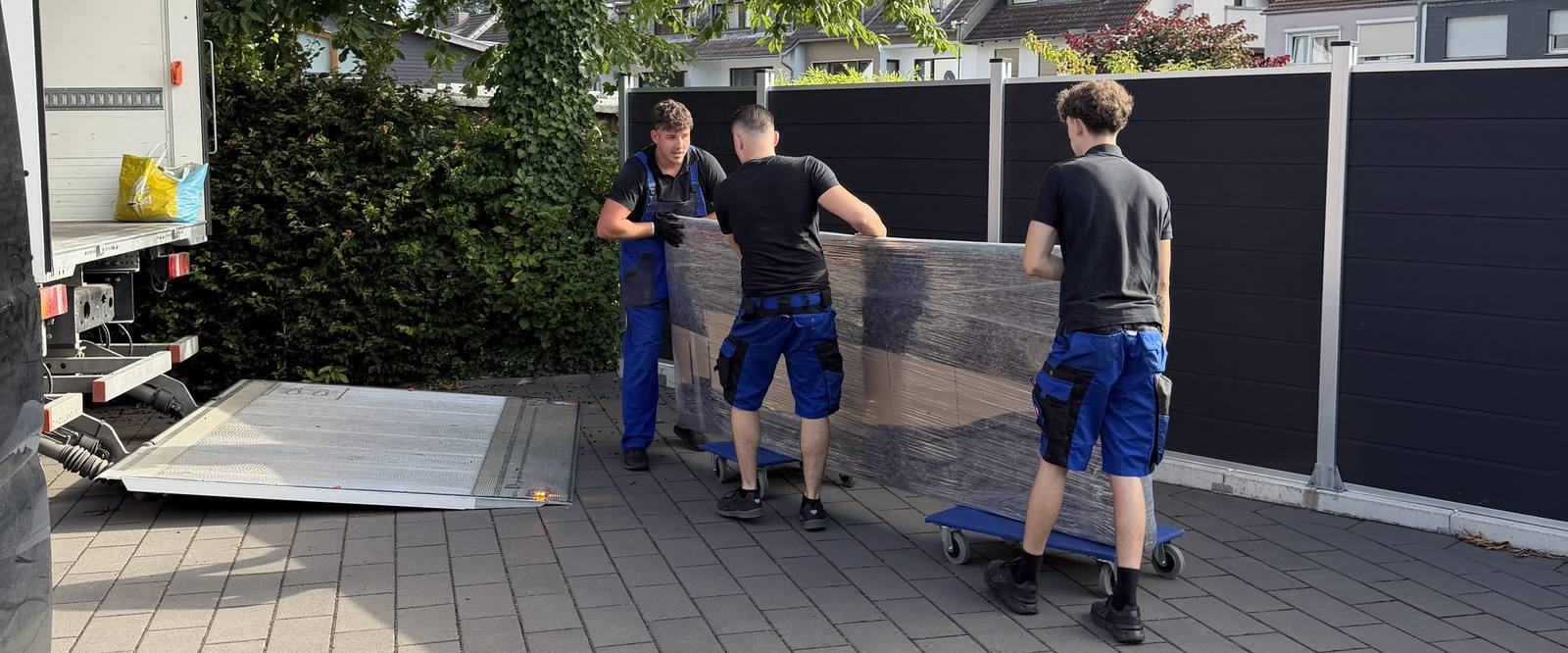 Umzugsunternehmen Leverkusen: Drei Mitarbeiter schieben ein foliertes Möbelstück auf Rollbrettern über einen Hof zum Umzugswagen, dessen Ladebordwand abgesenkt ist