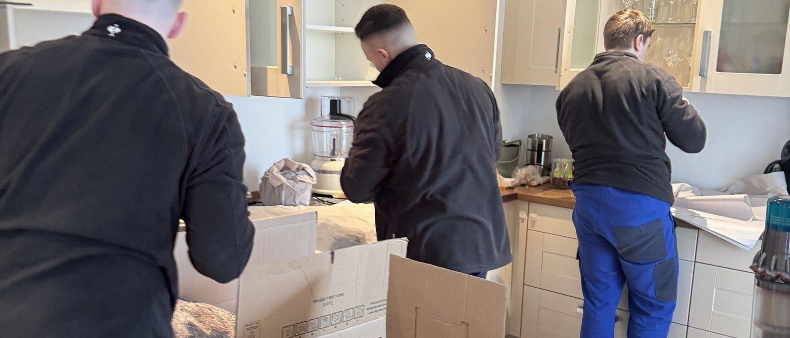 Umzugskartons und Kleiderboxen für Ihren Umzug in Leverkusen: Drei Mitarbeiter packen in einer Küche mit hellen Fronten Geschirr und Kleingeräte in Umzugskartons auf der Arbeitsplatte, in der Mitte ein offener Karton mit Packpapier.