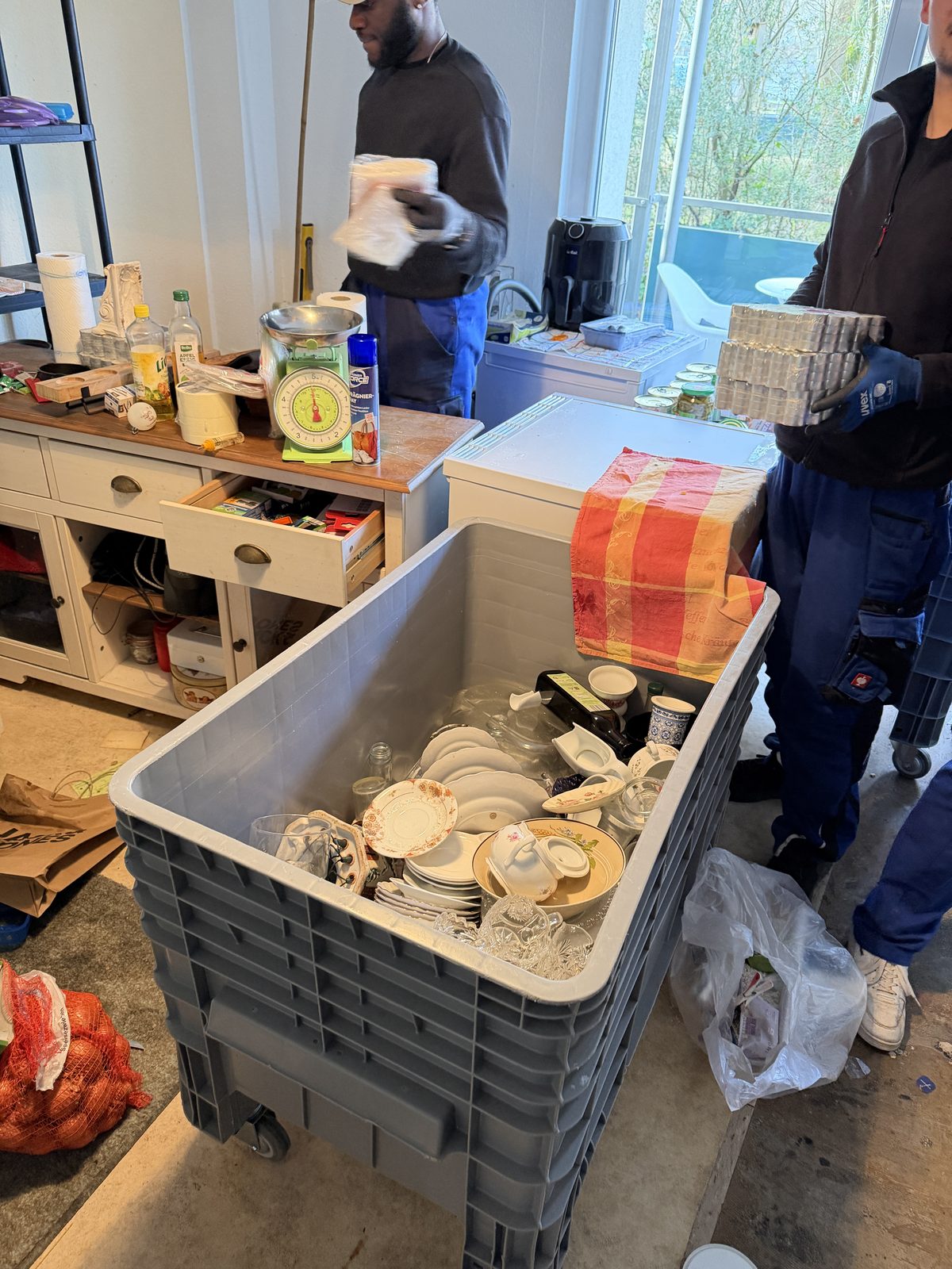 Zwei Mitarbeiter legen Geschirr und anderen Hausrat sortiert in einen großen grauen Rollbehälter.