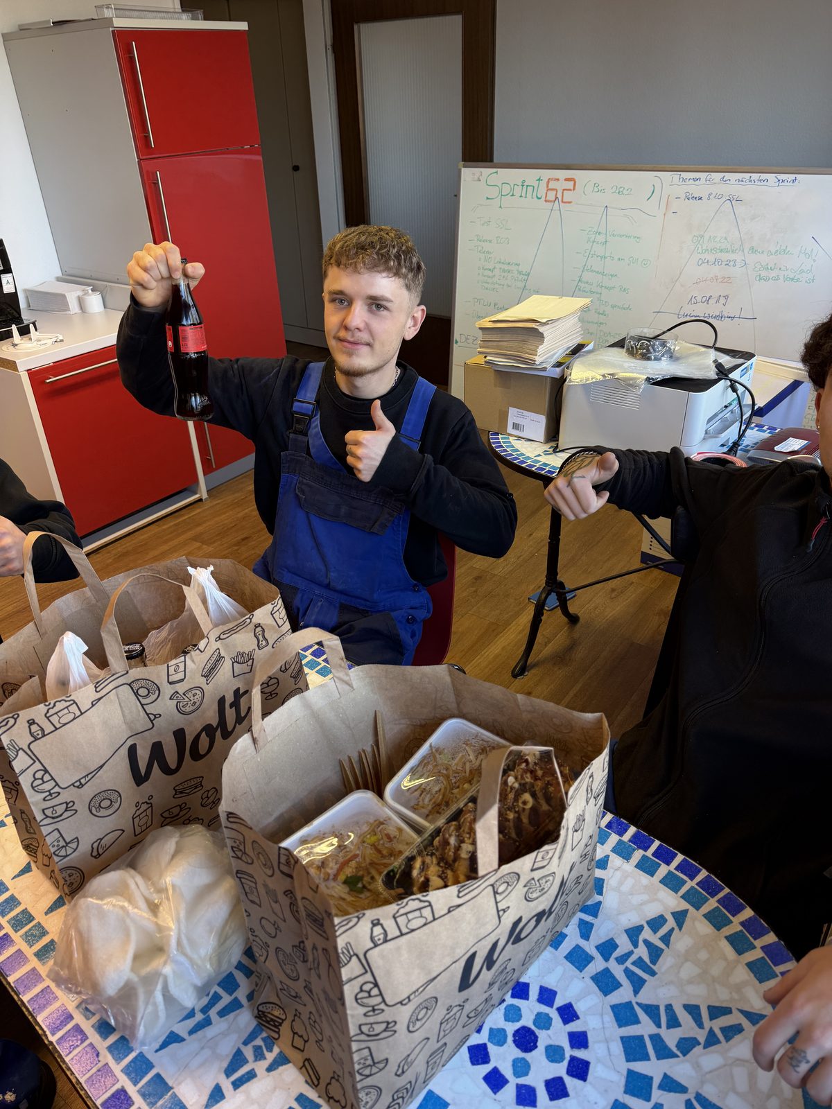 Ein Mitarbeiter hält beim Mittagessen mit den Kollegen ein Getränk in der Hand und hebt den Daumen.
