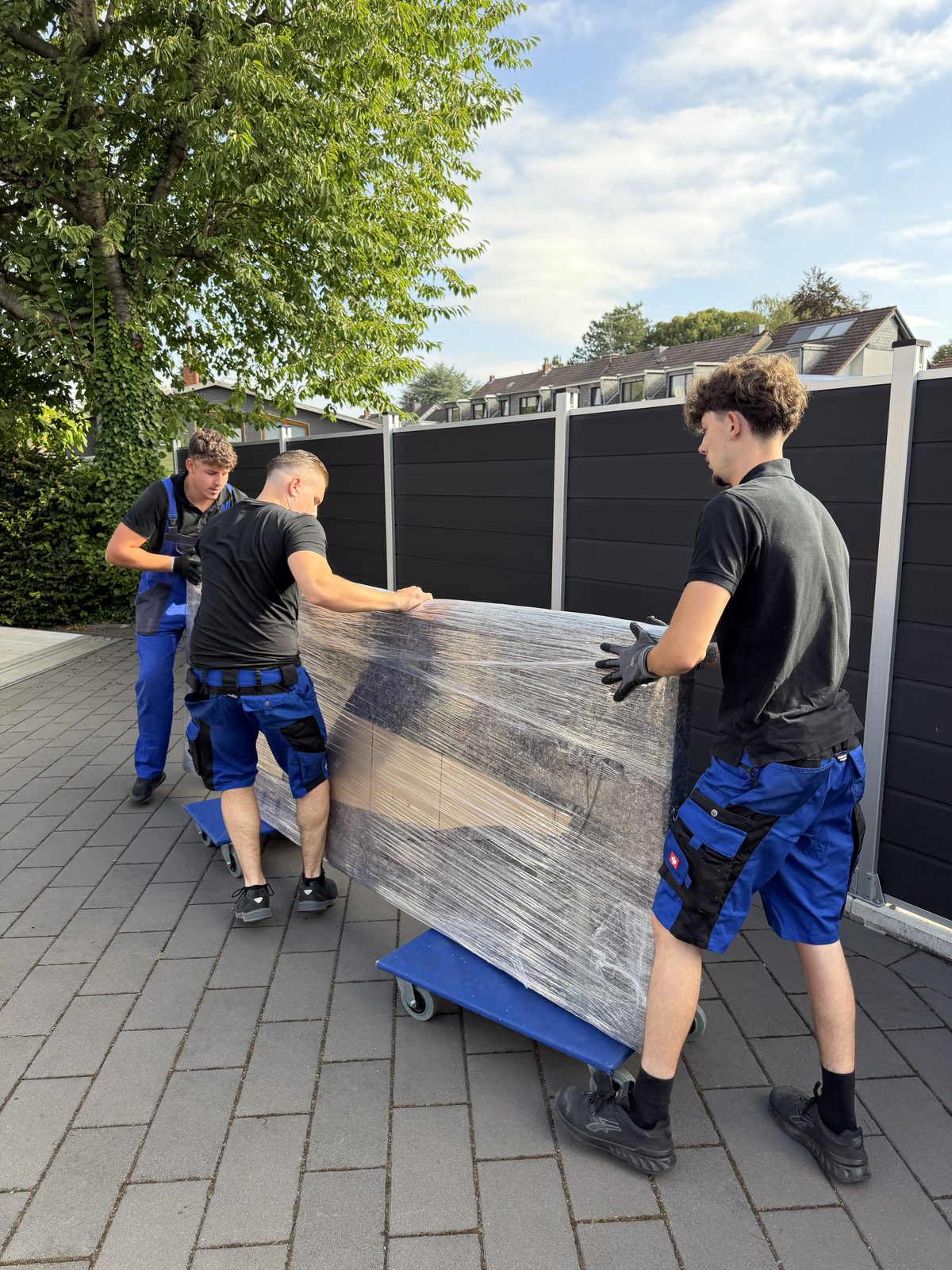 Drei Mitarbeiter rollen ein in Folie verpacktes Möbelstück auf Rollbrettern über die Einfahrt bis zum Umzugswagen.