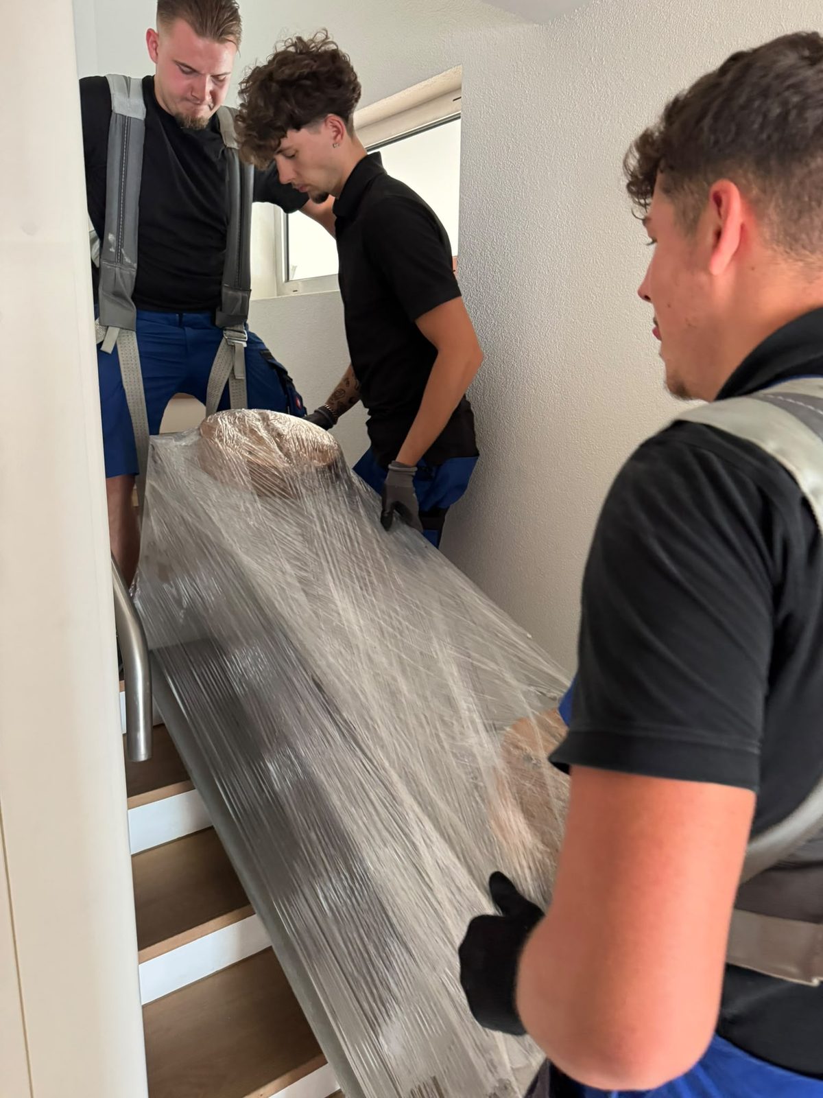 Drei Mitarbeiter tragen zusammen ein foliertes Möbelstück eine Treppe nach unten.