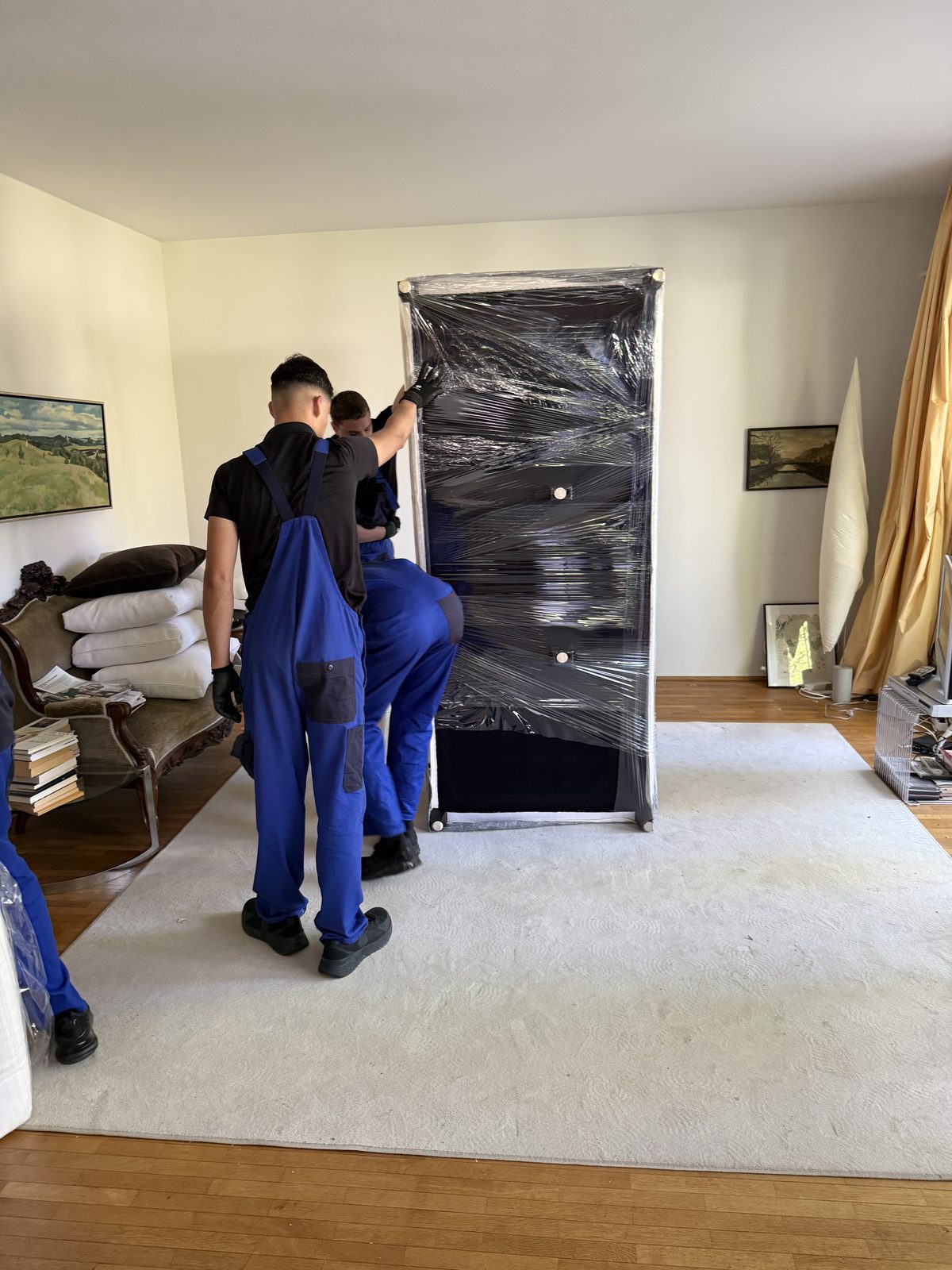 Zwei Mitarbeiter wickeln einen hohen Schrank in dunkle Folie ein, bevor er zum Wagen getragen wird.