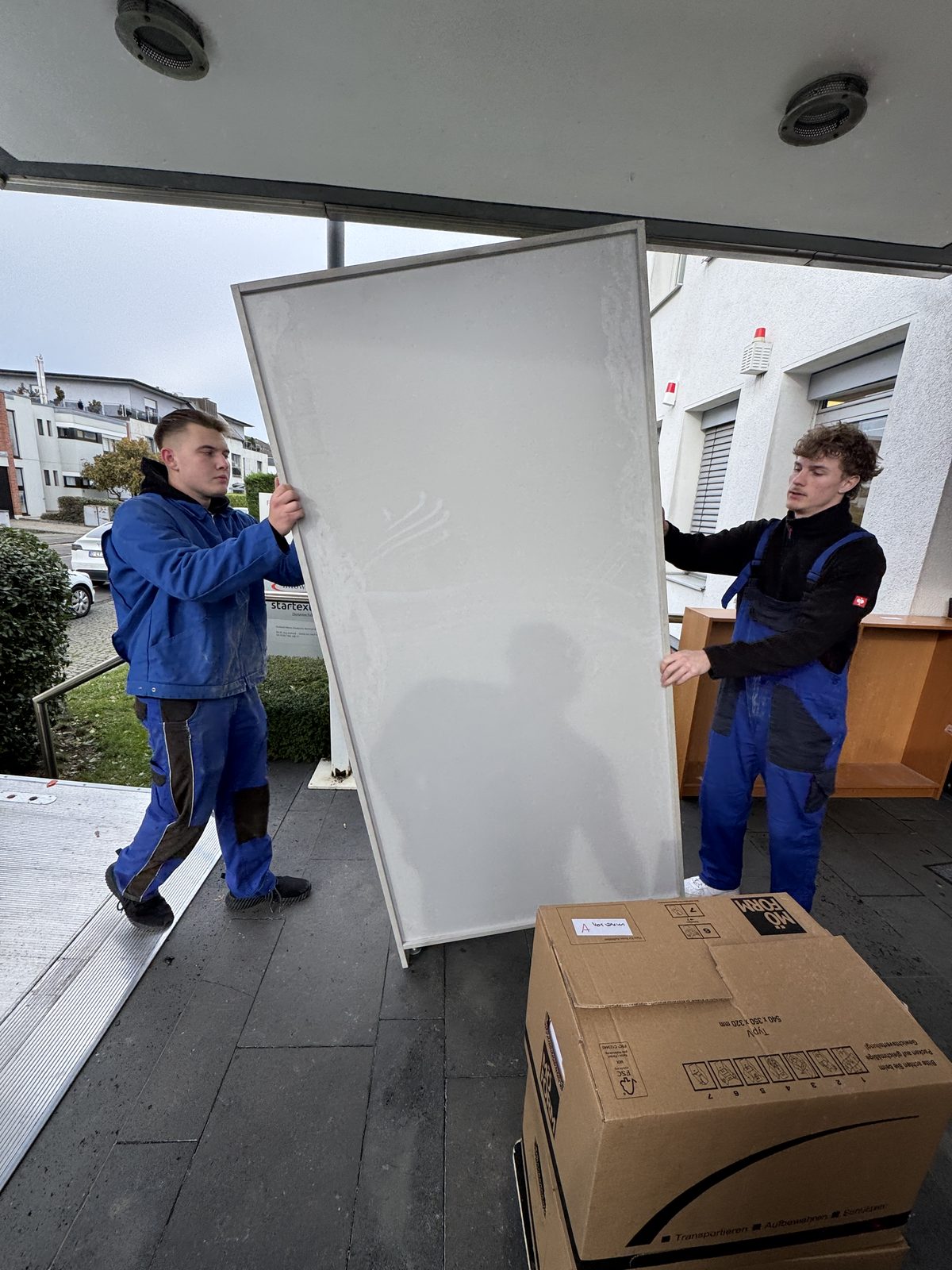 Zwei Mitarbeiter bringen eine große weiße Platte hochkant zum Umzugsfahrzeug.