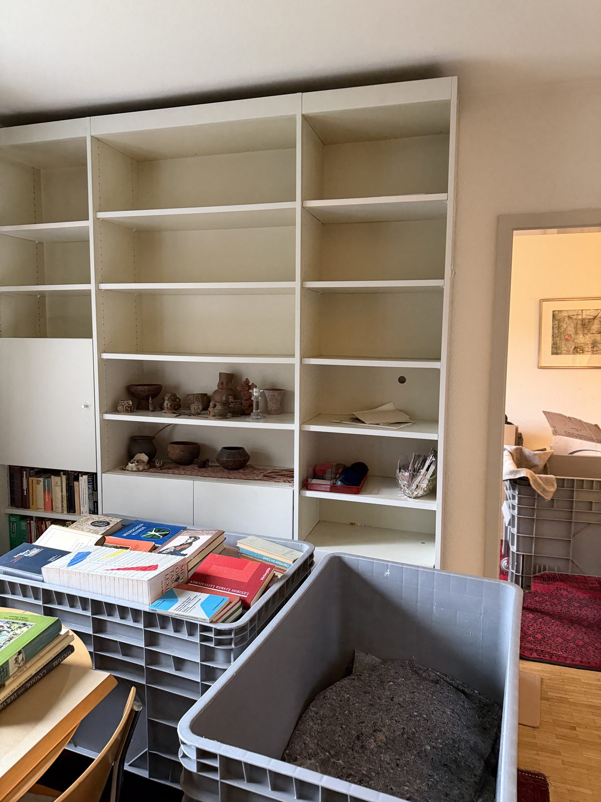 Ein Wandregal ist fast leer, nur wenige Dinge sind übrig, davor stehen Behälter voller Bücher.