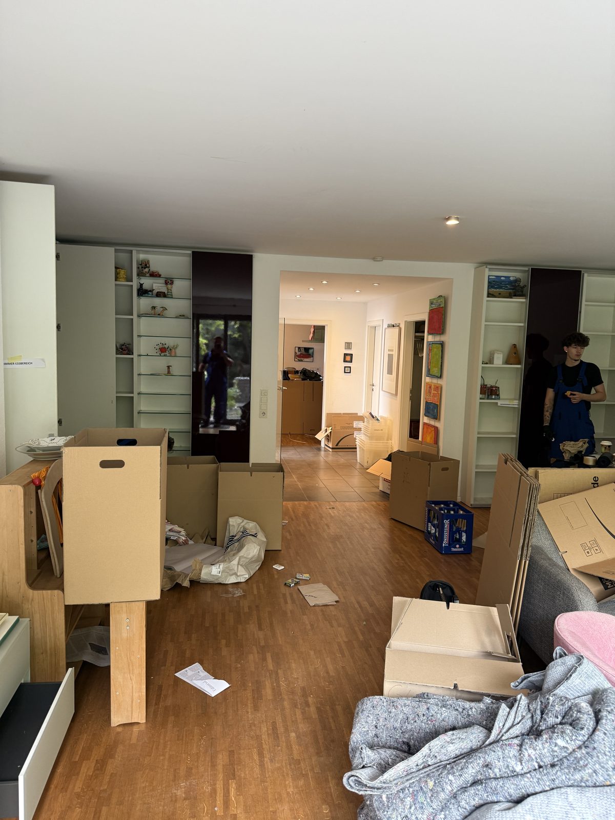 Ein Wohnzimmer mit ausgeräumten Einbauregalen und bereitgestellten Kartons, hinten arbeiten zwei Mitarbeiter.