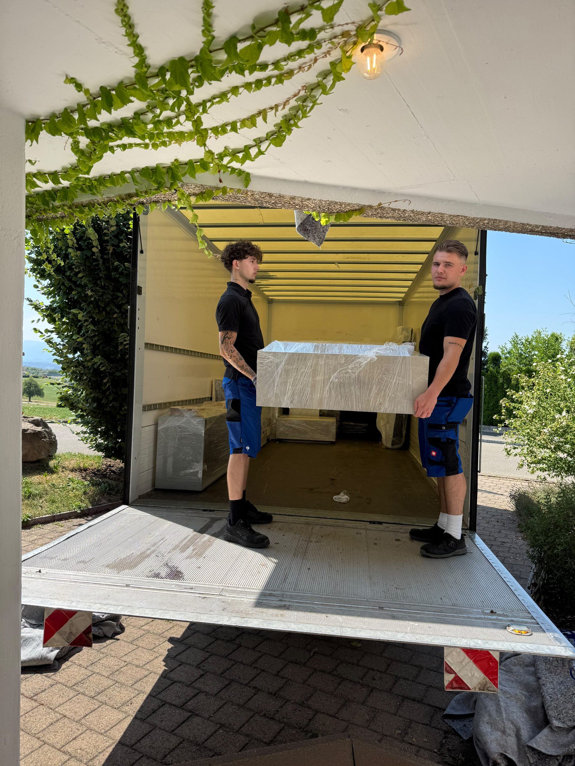 Zwei Mitarbeiter tragen ein foliertes Möbelstück über die Ladebordwand hinein in den Umzugswagen.